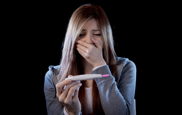 young sad woman scared and shocked holding pregnancy test positive result looking unhappy and in depression isolated on black background in unexpected pregnant woman concept